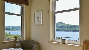 Un dormitorio con cama, una silla y ventana con vistas a un lago y colinas.