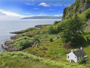 A white house with a stone roof, situated on a grassy hillside with a view of the sea and a distant mountain range.