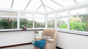 Une véranda avec un seul fauteuil beige, une petite table ornée d’un objet décoratif et une vue sur la verdure à l’extérieur.