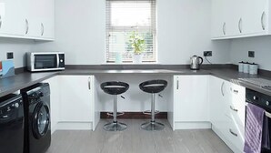 Une cuisine moderne avec des meubles blancs, un four à micro-ondes, un lave-linge, une bouilloire et deux tabourets de bar.