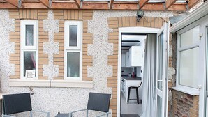 Une terrasse avec deux chaises et une table, une cuisine visible à travers une porte ouverte, et un mur de briques avec des fenêtres.