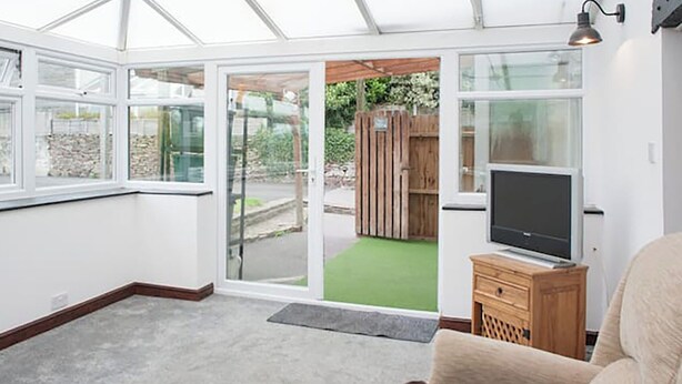 Une véranda équipée d’un téléviseur et de meubles en bois, avec un tapis vert et une vue sur un jardin.