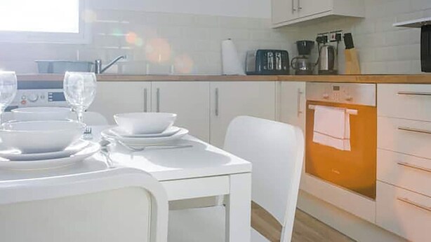 A modern kitchen with white cabinetry, a dining table set with white plates and glasses, and a clock on the wall. - Shirehampton Apartment (Shirehampton)