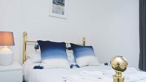 A neatly made bed with white linens and blue pillows, a bedside lamp, and a gold-framed headboard. - Shirehampton Apartment (Shirehampton)