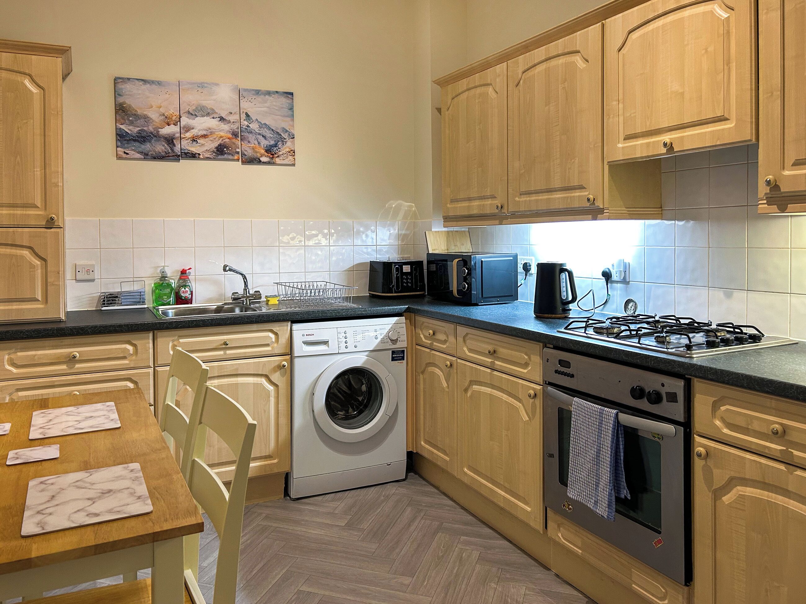 A modern kitchen with a Bosch washing machine, a microwave, a gas stove, and wooden cabinets.