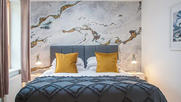 Une chambre avec un mur décoré d’un motif imitant le marbre, une tête de lit sombre et deux lampes de chevet.