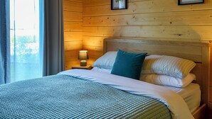 Un dormitorio estilo cabaña de madera con cama, almohadas, mesita de noche con lámpara y hojas enmarcadas en la pared.