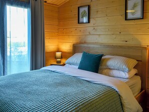 A wooden cabin-style bedroom with a bed, pillows, a nightstand with a lamp, and framed leaves on the wall. - Evergreen Lodge at Kingfisher Lakes (Brandesburton)