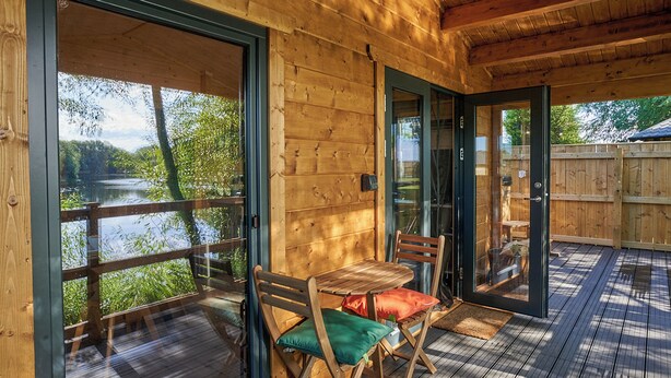 Una terraza de madera con una mesa y sillas, un ventanal de vidrio que da al exterior y vista a un lago.