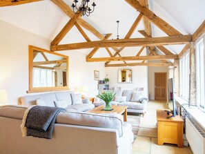Een ruime woonkamer met hoge plafonds en zichtbare houten balken, met twee banken, een salontafel en een televisie op een houten dressoir.