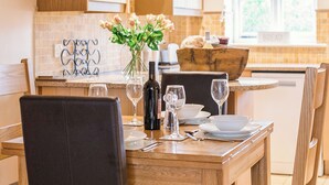 Una mesa de comedor lista con platos, vasos y una botella, rodeada de sillas en una cocina con armarios de madera y una ventana.