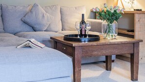 Un salón con un sofá gris, una mesa de centro de madera con una botella de vino y copas, una lámpara y un jarrón con flores.