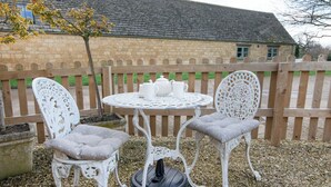 A patio with two white metal chairs and a table, surrounded by a wooden fence and a stone building in the background. - 1 bedroom accommodation in Chipping Campden (Chipping Campden)