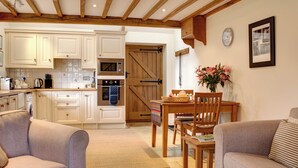 A cozy living room with a sofa, a coffee table with a book and a mug, a dining area with a wooden table and chairs, and a kitchen with white cabinets and wooden beams. - 1 bedroom accommodation in Chipping Campden (Chipping Campden)