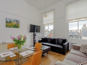 A modern living room with a glass dining table, leather sofa, and a large window with a city view.