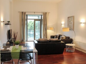 Ein modernes Wohnzimmer mit einer Couch, einem Esstisch und Stühlen. Ein großes Fenster mit Vorhängen, eine Stehlampe und eine kleine Pflanze auf einem Regal.