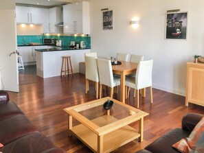 A modern living room with a glass coffee table, a dining area with a wooden table and white chairs, a kitchen with white cabinets and a green backsplash, and a small island with a microwave and toaster. - Crown Apartments 3B - UK3385 (Newcastle upon Tyne)