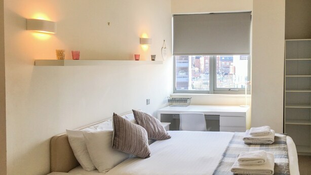 Ein Schlafzimmer mit Bett, Nachttischen, einem Schreibtisch und einem Fenster mit Blick auf Gebäude.