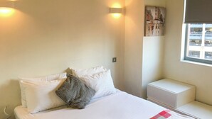 Ein Hotelzimmer mit einem Bett, Kissen, einem Nachttisch, einem Fenster und einem an der Wand hängenden Kunstwerk.