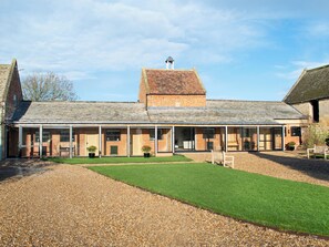 A large, single-story brick building with a sloped roof, a covered outdoor area with seating, and a gravel driveway. - The ultimate chic space for those seeking a stylish getaway. (Chipping Campden)