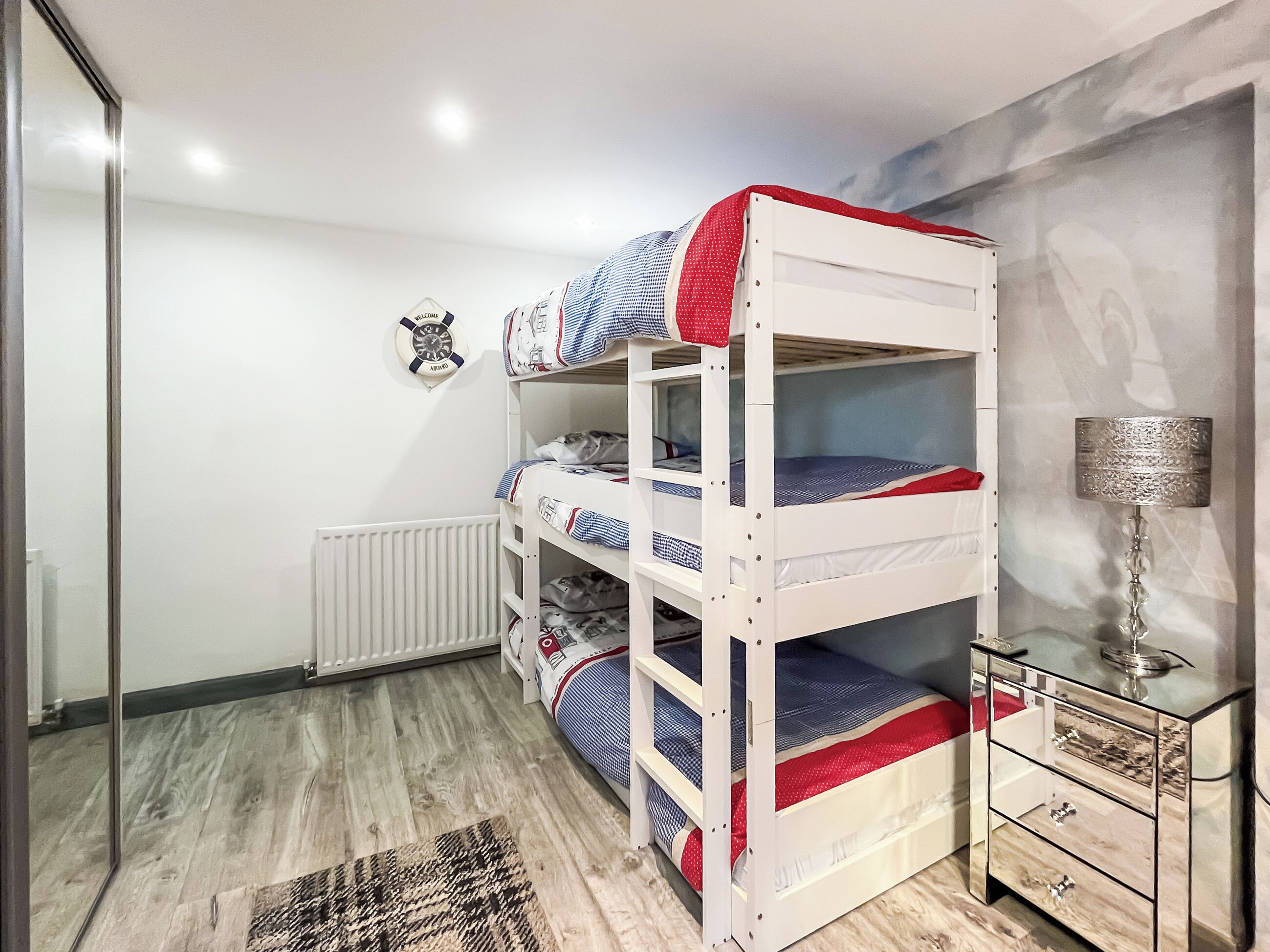 A bunk bed room with a radiator, a small table with drawers, and a wall-mounted lamp.
