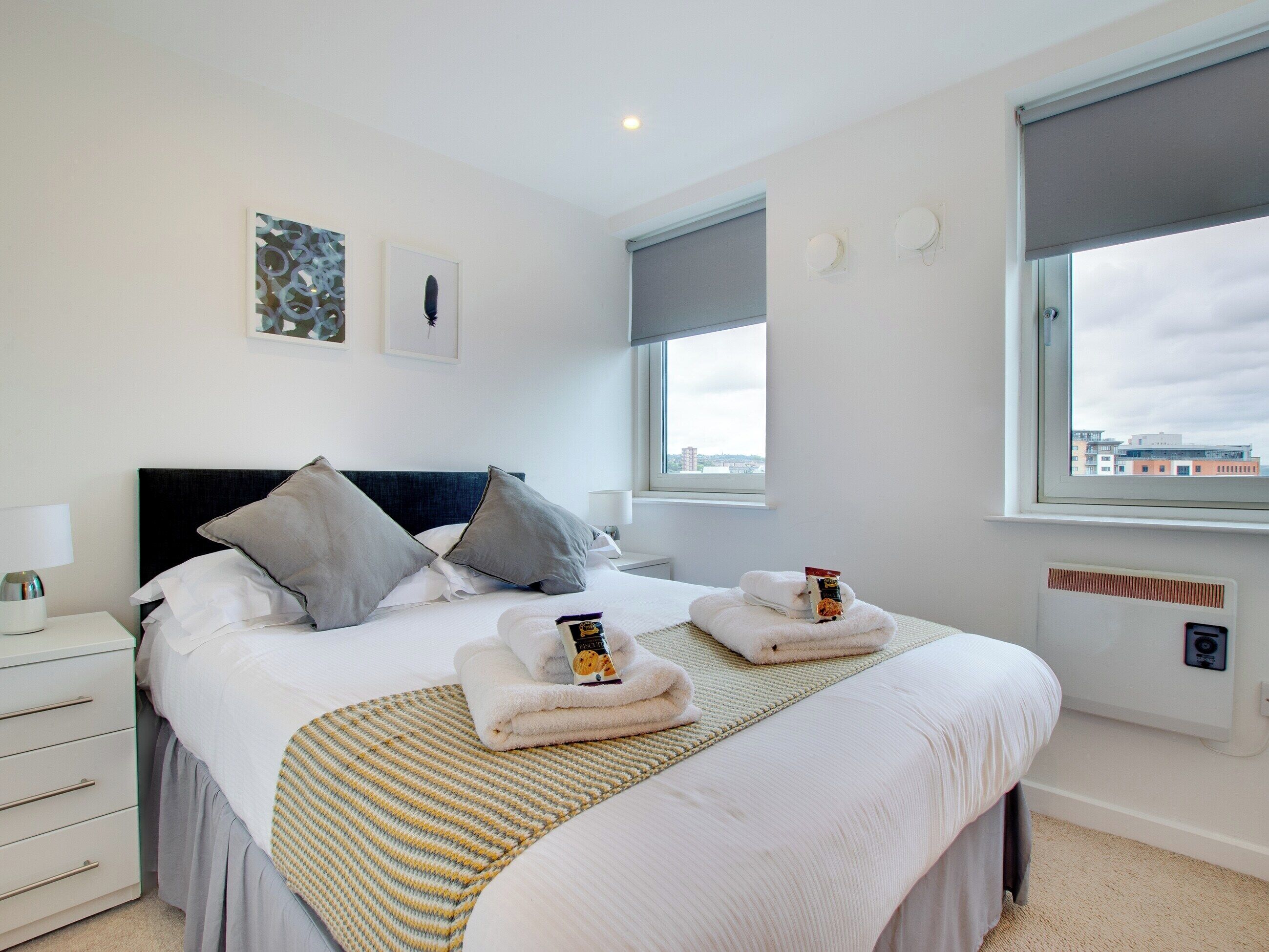 A neatly made bed with pillows, a bedside table with a lamp, and a window with a view of buildings.