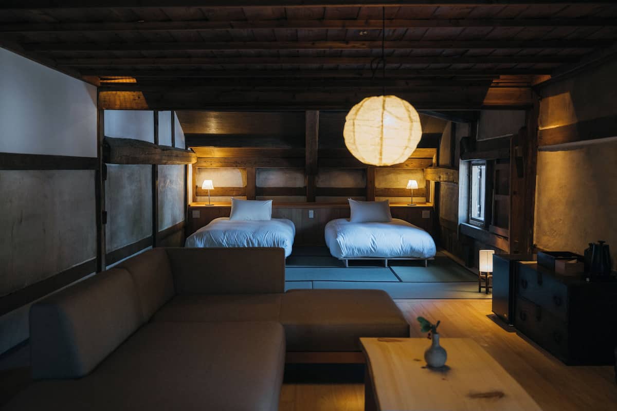 Ein traditioneller japanischer Raum mit Tatami-Boden, zwei Betten, einer niedrigen Holzbalkendecke und einer hängenden Leuchte.