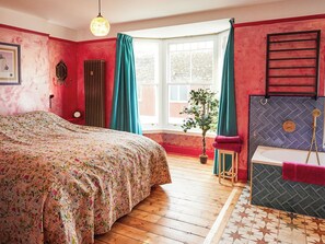 A bedroom with a floral bedspread, a window with curtains, a potted plant, and a bathtub with a shower head. - 4 bedroom accommodation in Combe Martin, near Ilfracombe (Combe Martin, near Ilfracombe)