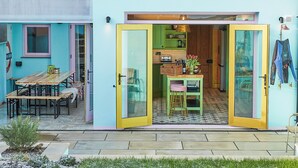 Un bâtiment d’un bleu vif, avec une porte jaune qui donne sur une cuisine équipée de meubles verts et une petite table à manger entourée de chaises.