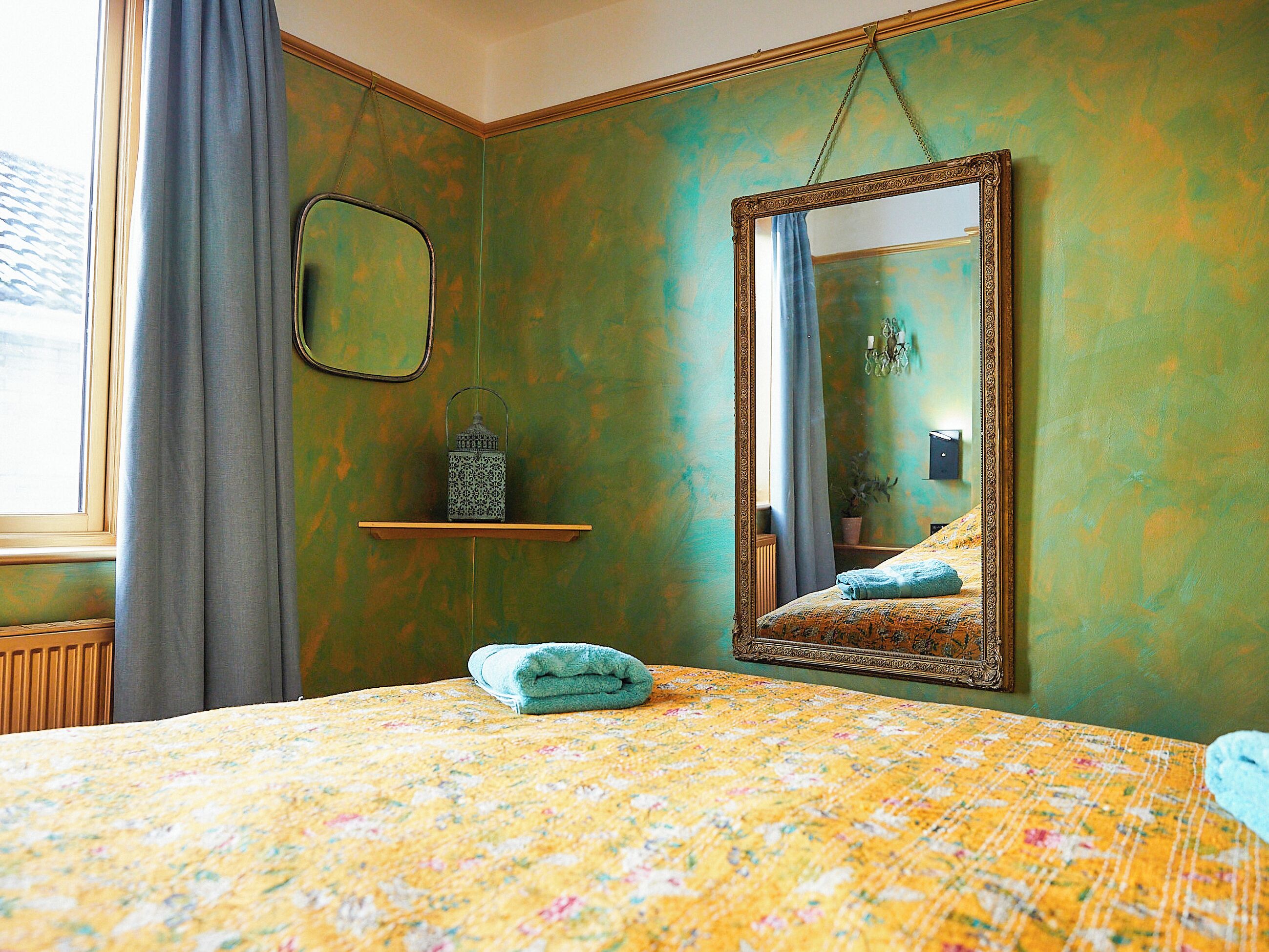 A room with a bed, a mirror, a shelf with a lantern, and a window with curtains.