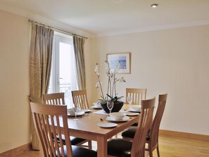 Une salle à manger avec une table en bois dressée pour quatre personnes, entourée de chaises, avec une fenêtre et des rideaux d’un côté, et un tableau encadré au mur.