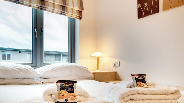 Een hotelkamer met een bed, nachtkastje, lamp en raam met uitzicht op gebouwen.