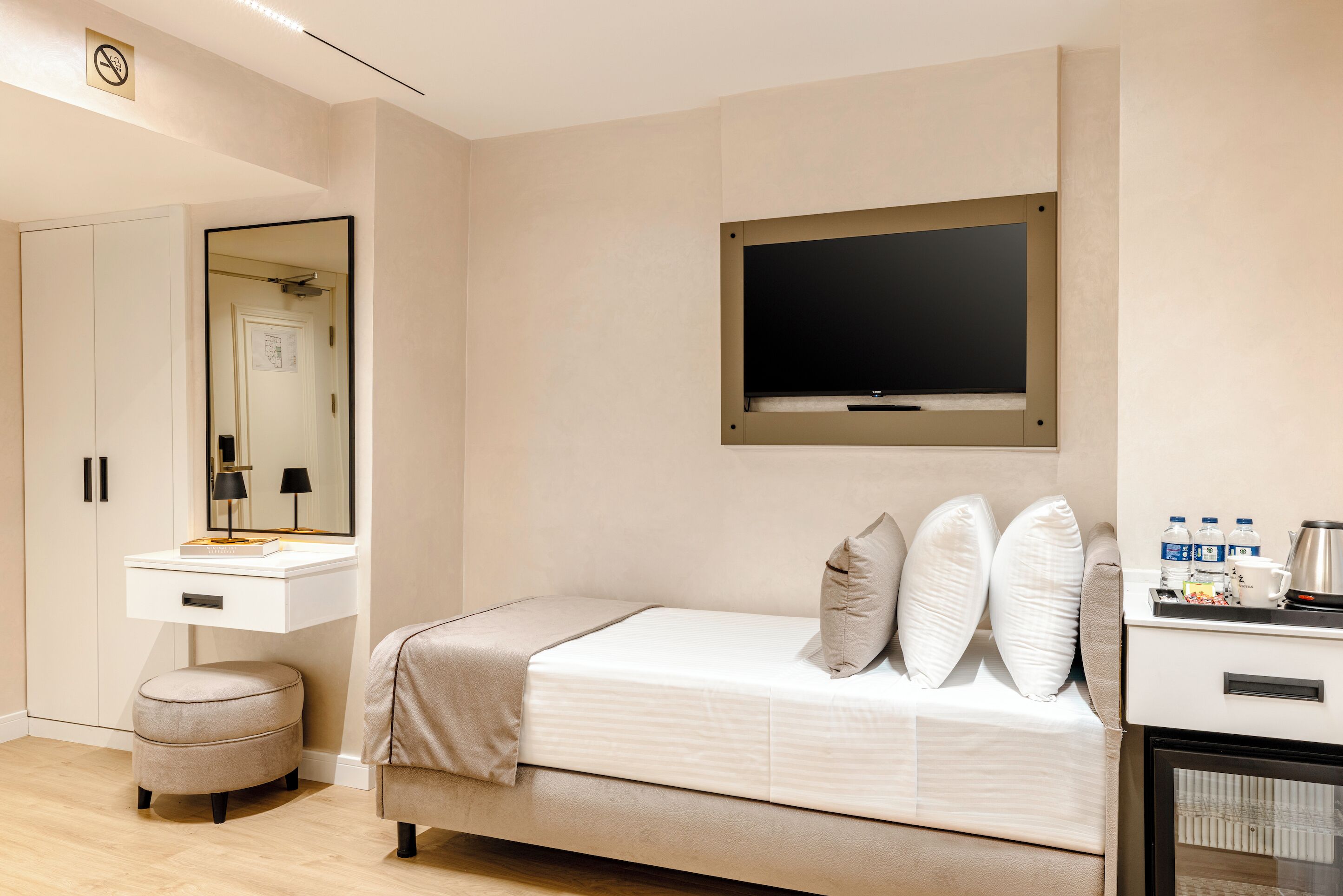 A bedroom with a bed, a vanity with a mirror, a television mounted on the wall, and a small kitchenette area.