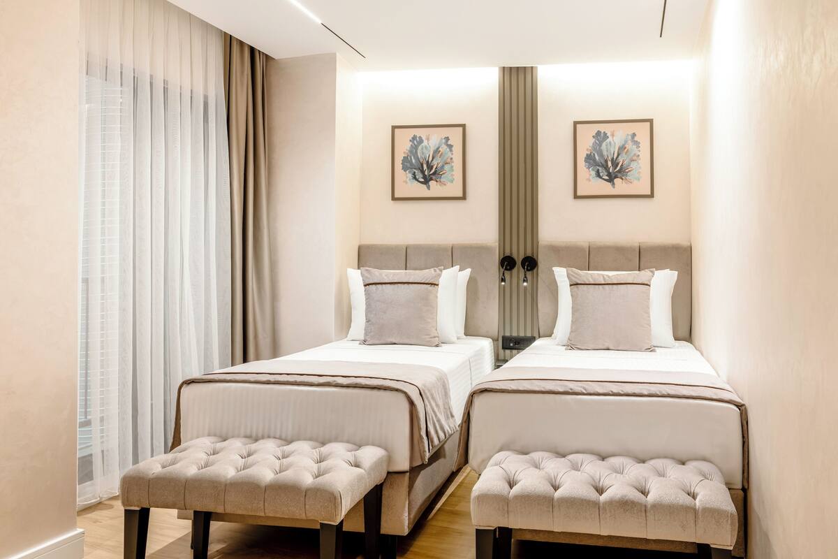 A hotel room with two beds, a large window with sheer curtains, and two wall-mounted lamps.