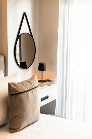 Ein ordentlich eingerichtetes Hotelzimmer mit Bett, Nachttischlampe, rundem Spiegel und einem Fenster mit durchsichtigen Vorhängen.