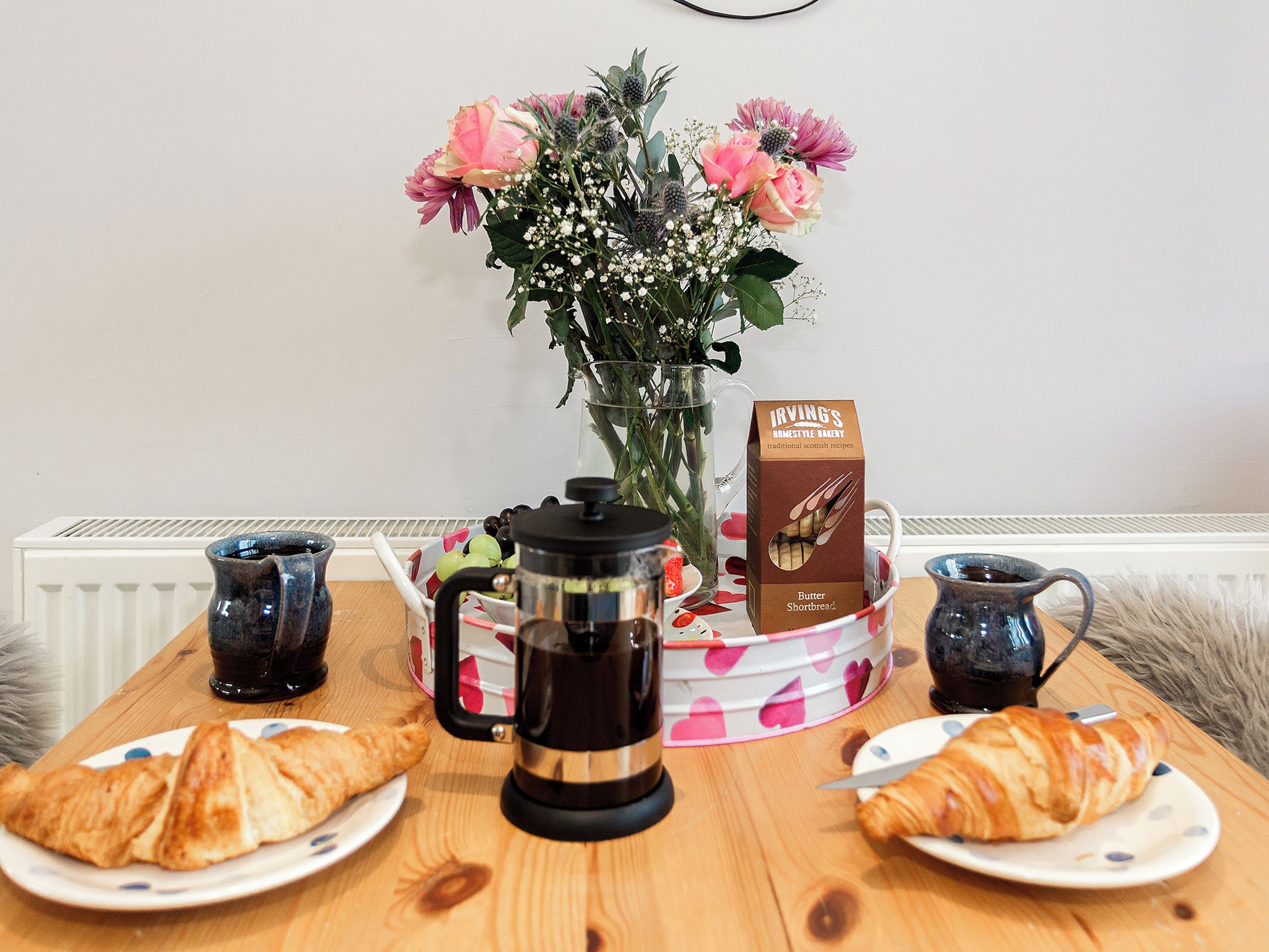 Una mesa de desayuno con croissants, una cafetera, una caja de galletas de mantequilla Irving's y un jarrón con flores.