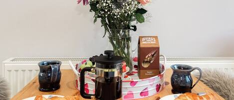 Una mesa de desayuno con croissants, una cafetera, una caja de galletas de mantequilla Irving's y un jarrón con flores.
