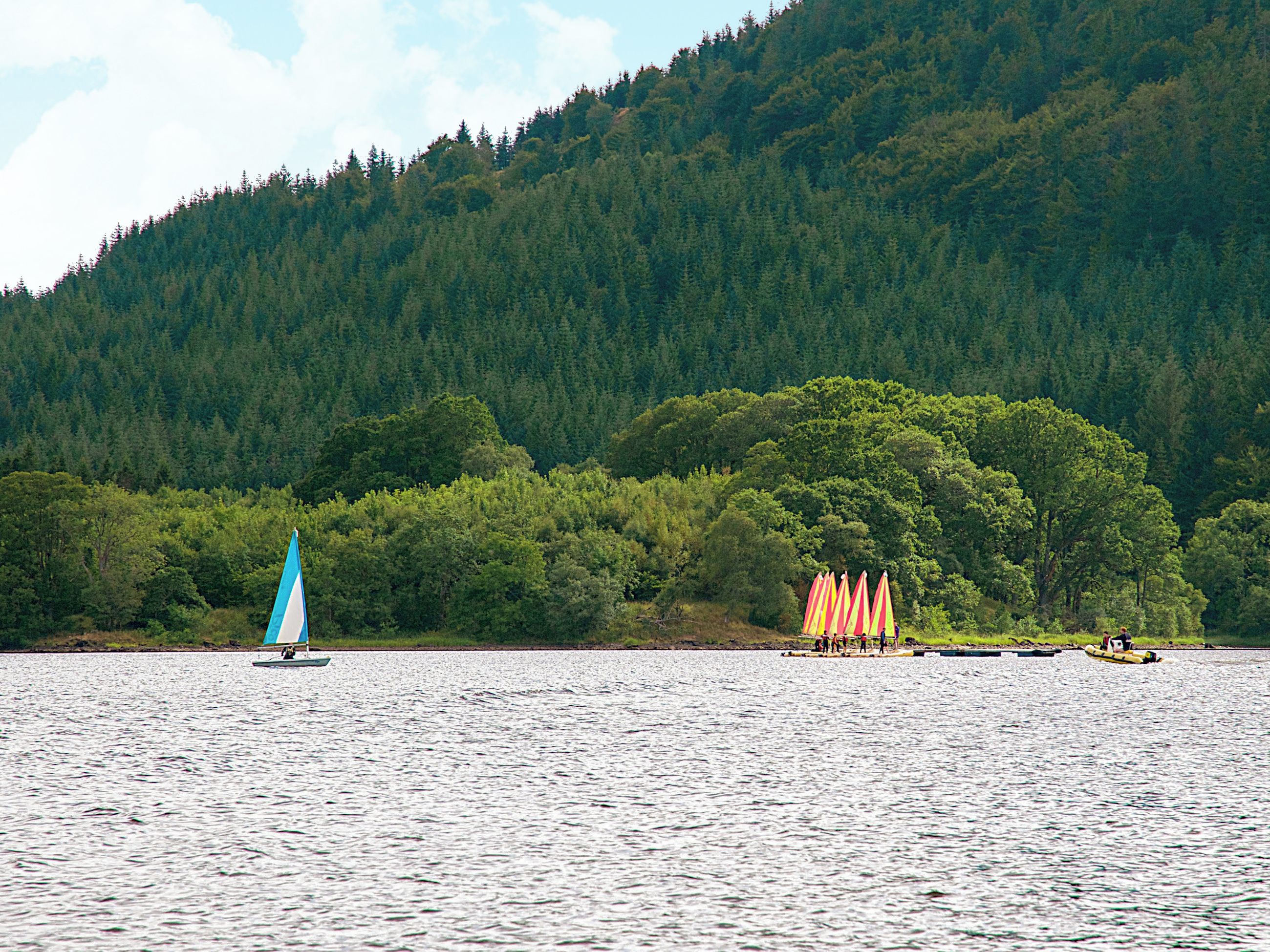 A lake with a sailboat and a group of colorful umbrellas on the shore.