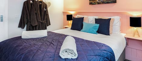 A neatly made bed with a blue quilt, a white towel, and a painting of a landscape on the wall.