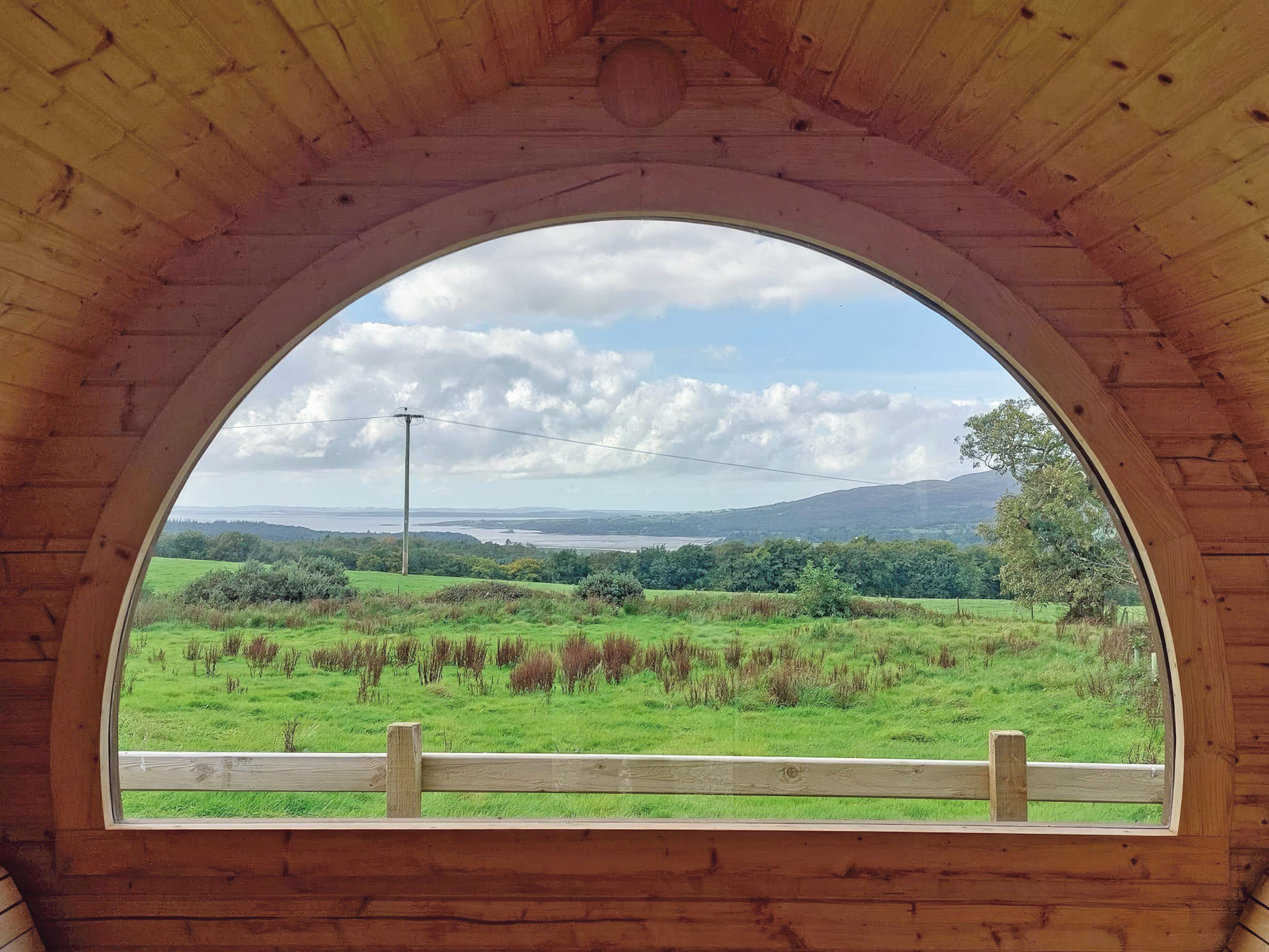 A wooden arched window with a view of a green field and distant hills.