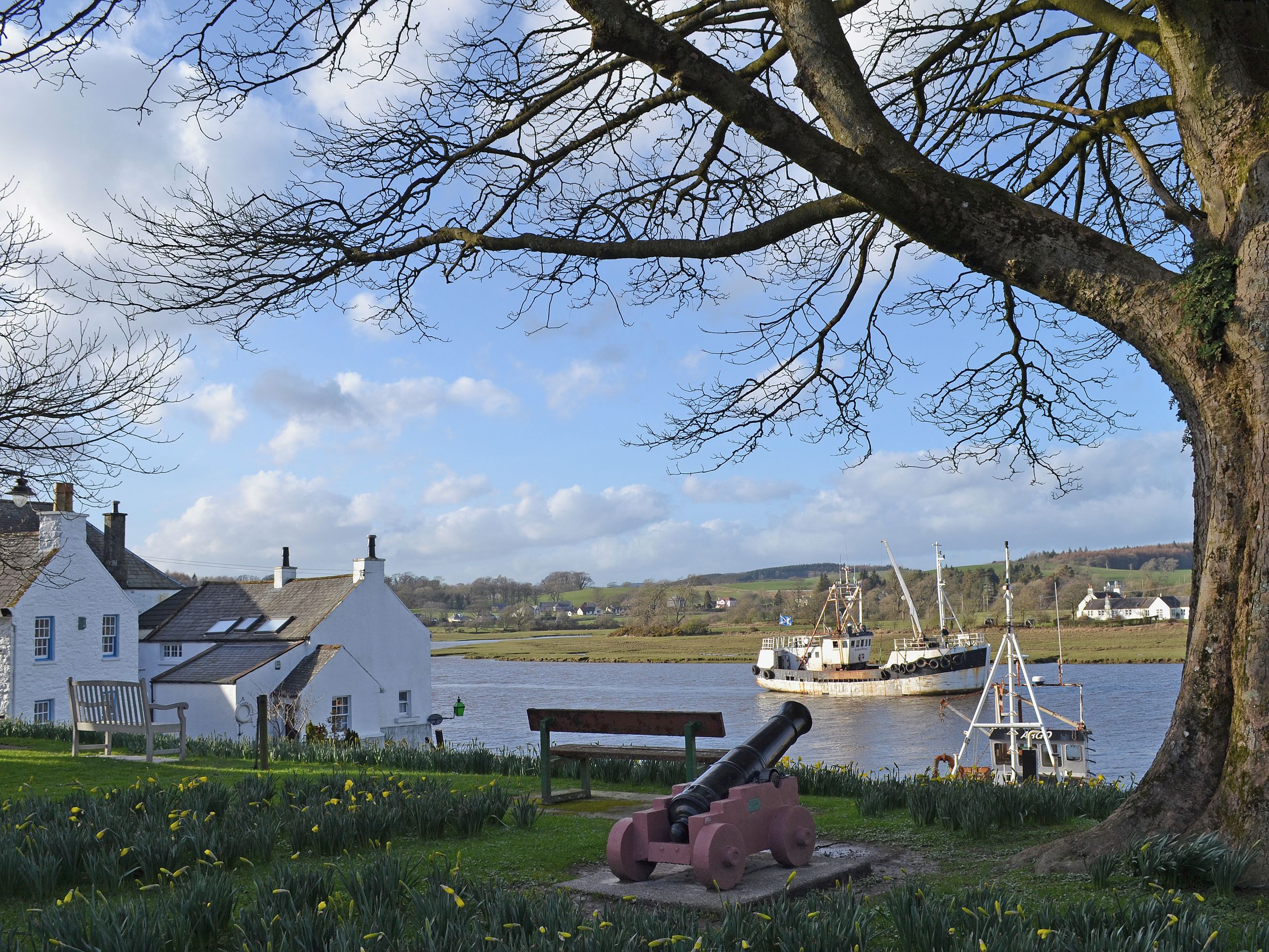 A white building with a sloped roof, a bench, and a cannon near a body of water.