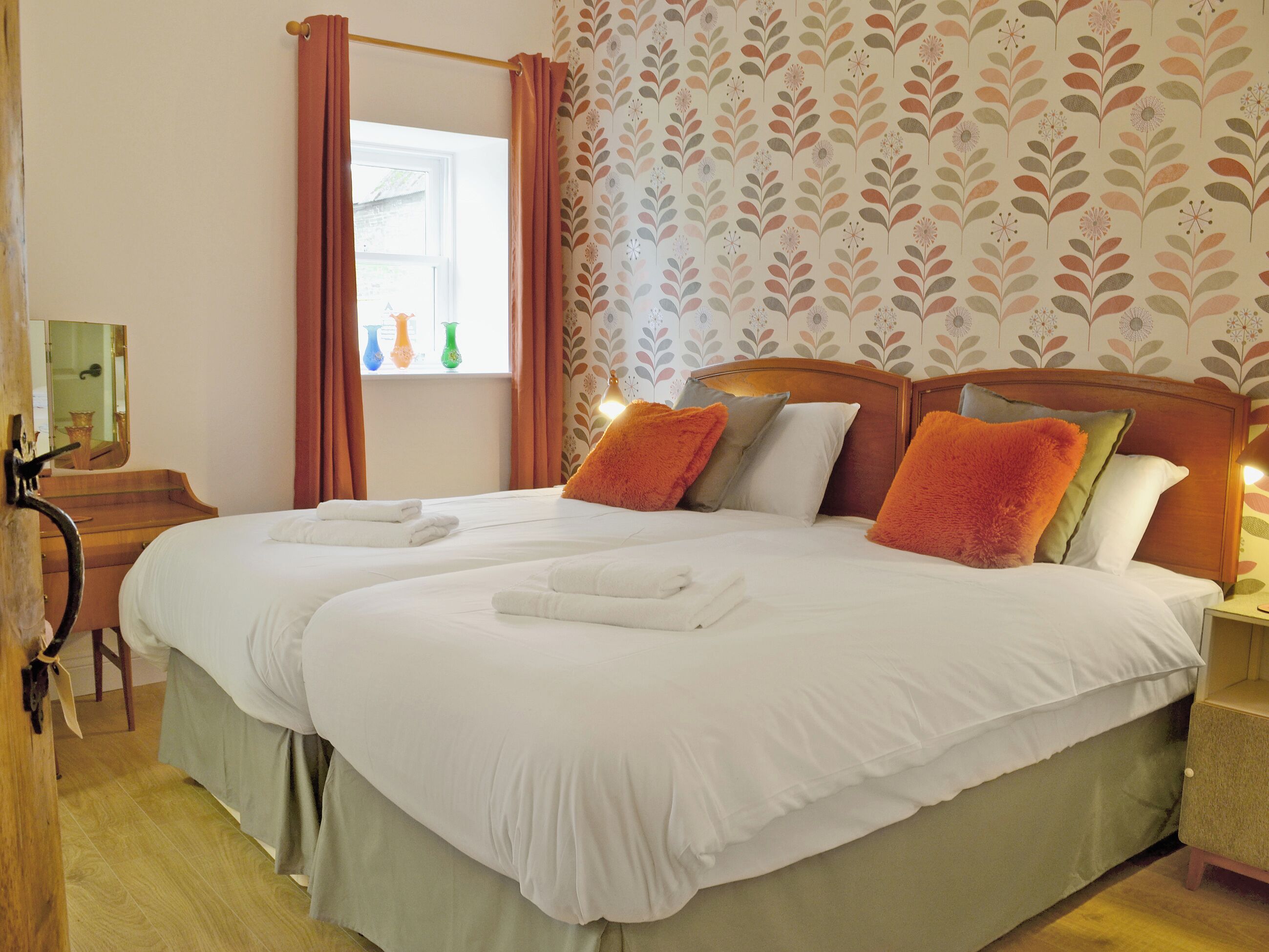 Cama doble con sábanas blancas, dos almohadas naranjas y cabecero de madera.