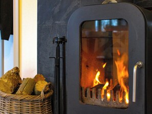 A wood-burning stove with visible flames, a metal handle, and a basket of firewood beside it. - 1 bedroom accommodation in Aberhafesp, near Newtown (Newtown)