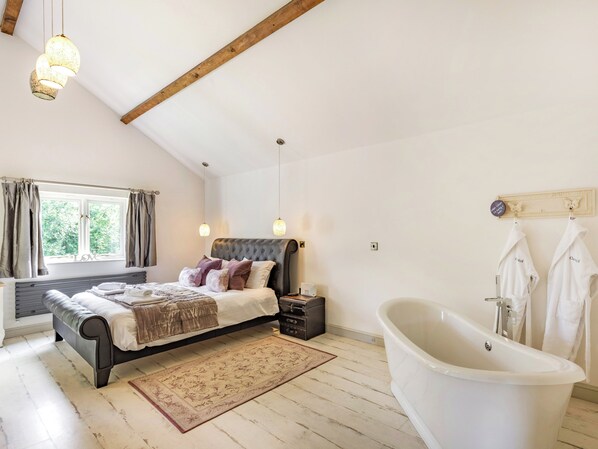 A bedroom with a bed, a bedside table, a nightstand, a window with curtains, a ceiling with exposed beams, a hanging light fixture, and a bathtub. - 1 bedroom accommodation in Aberhafesp, near Newtown (Newtown)