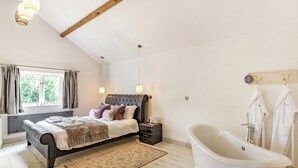 A bedroom with a bed, a bedside table, a nightstand, a window with curtains, a ceiling with exposed beams, a hanging light fixture, and a bathtub.