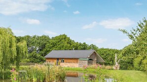 A rustic wooden cabin with a sloped roof, situated beside a pond with surrounding greenery and trees.