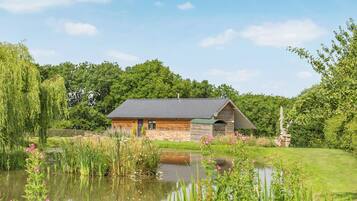 A rustic wooden cabin with a sloped roof, situated beside a pond with surrounding greenery and trees.