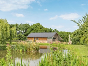 A rustic wooden cabin with a sloped roof, situated beside a pond with surrounding greenery and trees.