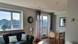 Ein Wohnzimmer mit einer Couch, einem Esstisch mit Stühlen, einer Lampe, einer Uhr und Blick auf Gebäude durch ein Fenster.