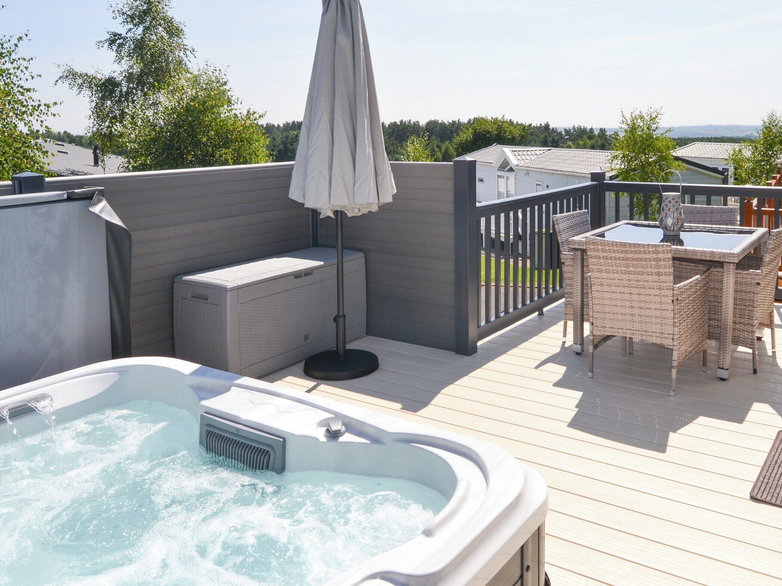 Ein Whirlpool auf einer Holzterrasse mit Tisch und Stühlen, umgeben von einem Holenzaun und Bäumen im Hintergrund.